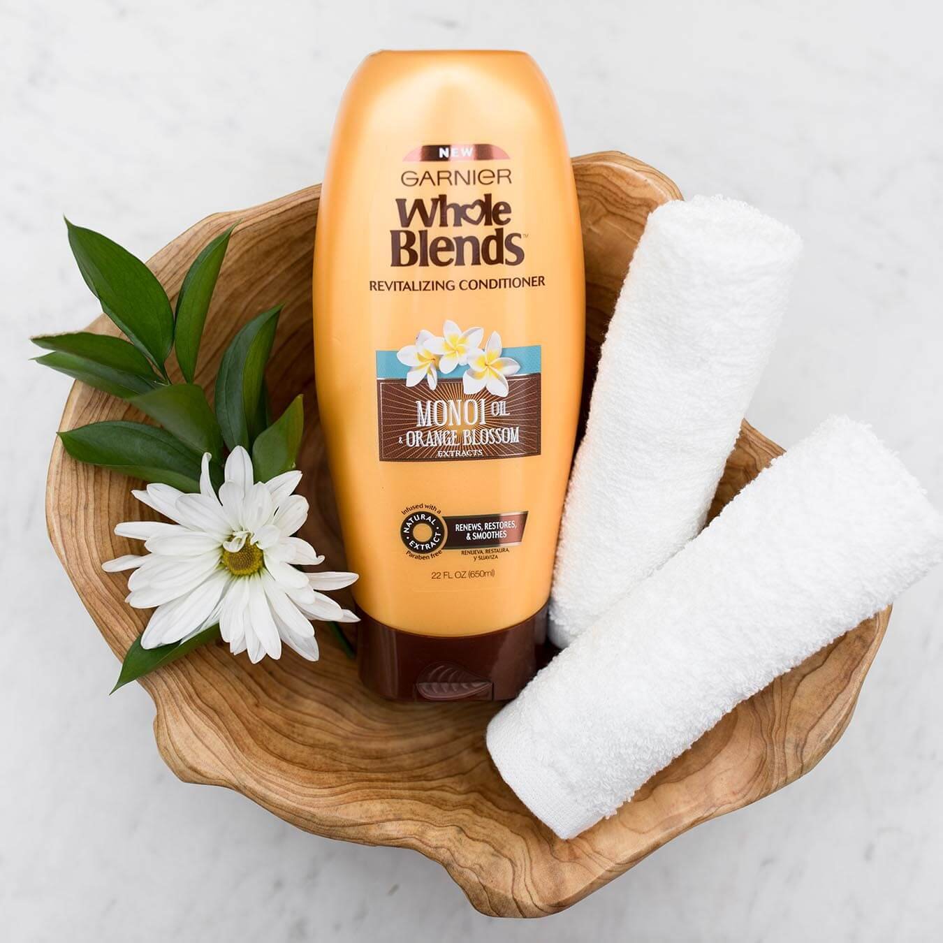 Whole Blends Monoi Oil Conditioner with Orange Blossom Extract in an irregular wooden bowl with a monoi blossom and leaves and two rolled white hand towels on white marble.